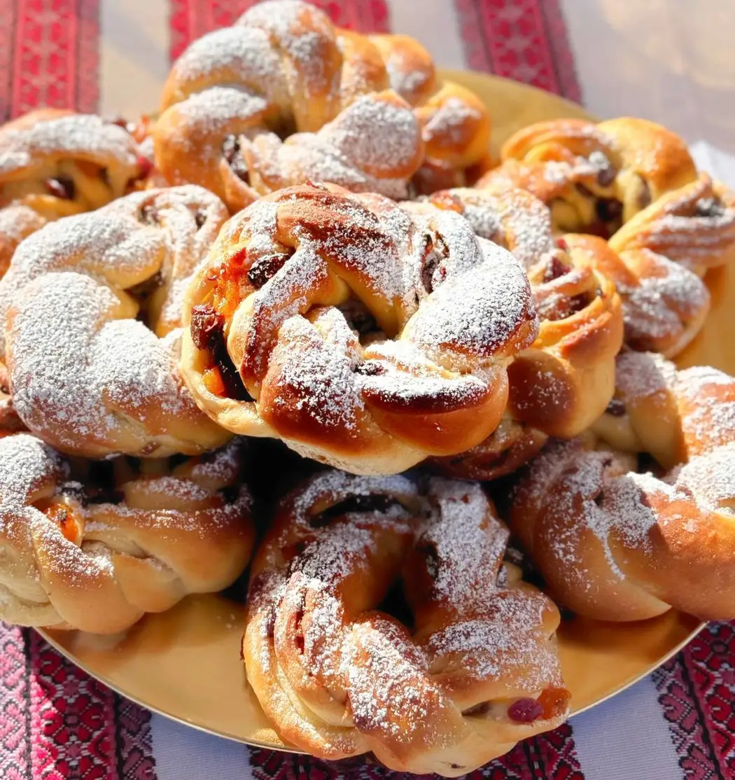 Braided sweet buns with dried fruit, dusted with icing sugar, on a gold plate over a red embroidered cloth